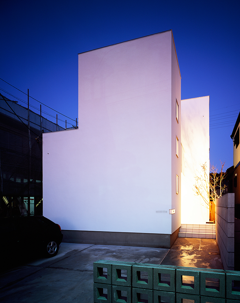 フラットで真っ白な外壁に、サイドから温かみのある光が溢れ出る夜の外観